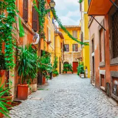 Urokliwa, brukowana uliczka w Trastevere, Rzym, zdominowana przez pomarańczowe i żółte fasady budynków, porośnięta bujną zielenią.