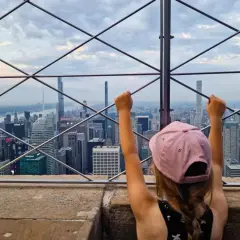 Dziecko z uniesionymi rękami podziwia panoramę Nowego Jorku z tarasu widokowego. Widać imponującą empire state building wysokość i inne drapacze chmur.