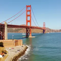 Słynny czerwony most w USA, Golden Gate, góruje nad błękitną wodą. Obok niego budynek z cegły i zielone drzewa.