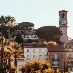 Słynny napis CANNES góruje nad malowniczym festiwalowym miastem na Lazurowym Wybrzeżu, z palmami i zabytkową wieżą zegarową w tle.