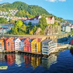 Kolorowe kamienice nad wodą, idealne na wycieczki do Norwegii. W tle zielone wzgórza i zabudowania.