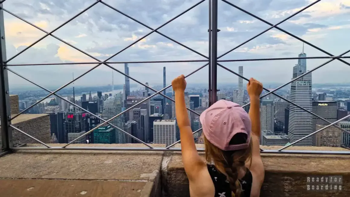 Dziecko z uniesionymi rękami podziwia panoramę Nowego Jorku z tarasu widokowego. Widać imponującą empire state building wysokość i inne drapacze chmur.