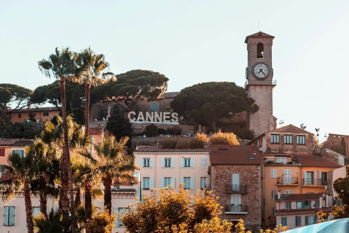 Słynny napis CANNES góruje nad malowniczym festiwalowym miastem na Lazurowym Wybrzeżu, z palmami i zabytkową wieżą zegarową w tle.