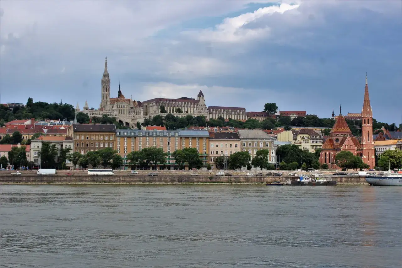 Widok na Dunaj i zabytkowe budynki w Budapeszcie. W oddali majestatyczny Zamek Królewski i Kościół Macieja, a bliżej ceglana wieża kościelna.