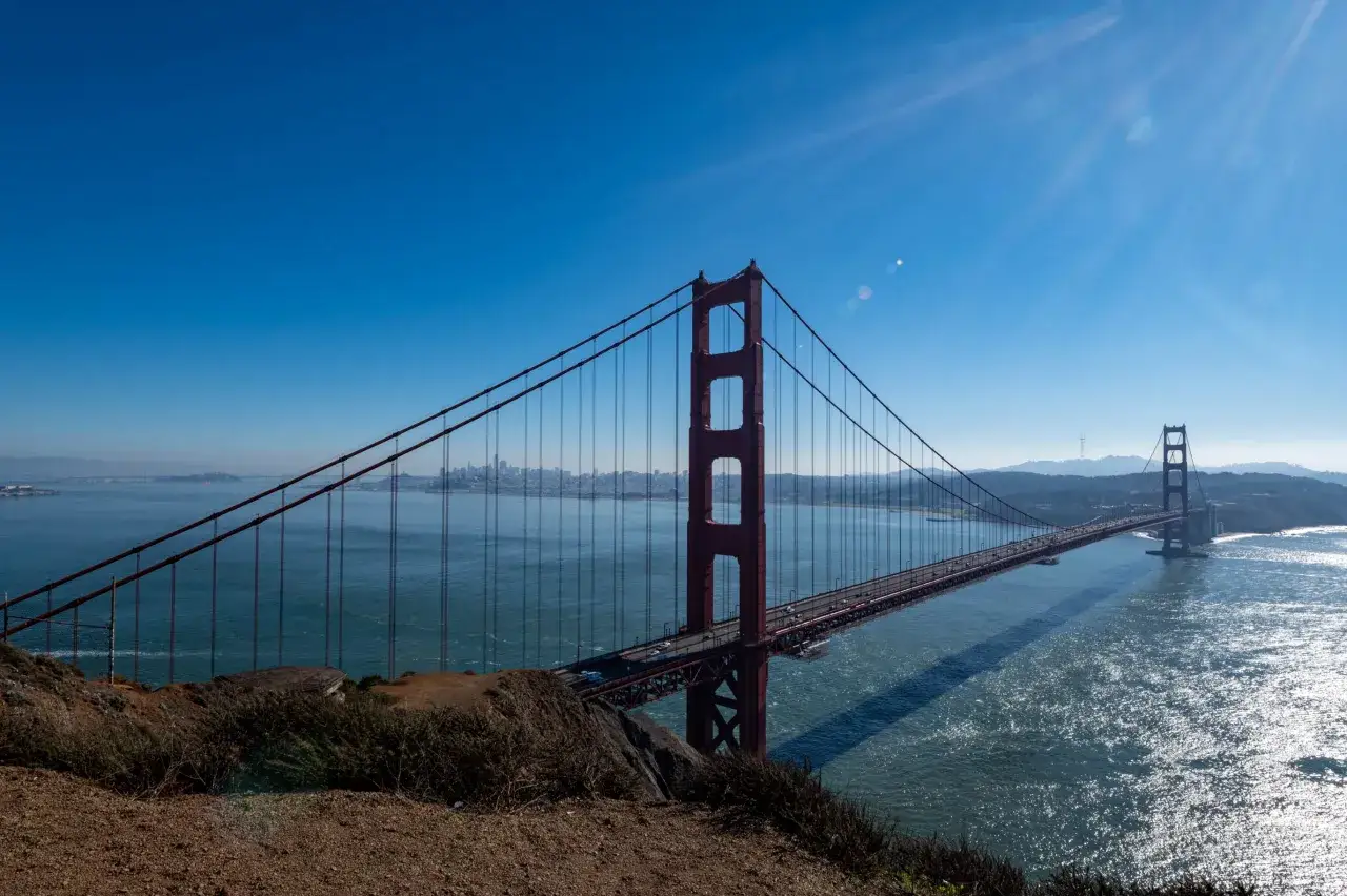 Słynny czerwony most w USA, Golden Gate, rozciąga się nad błękitną wodą. W oddali widać panoramę miasta.