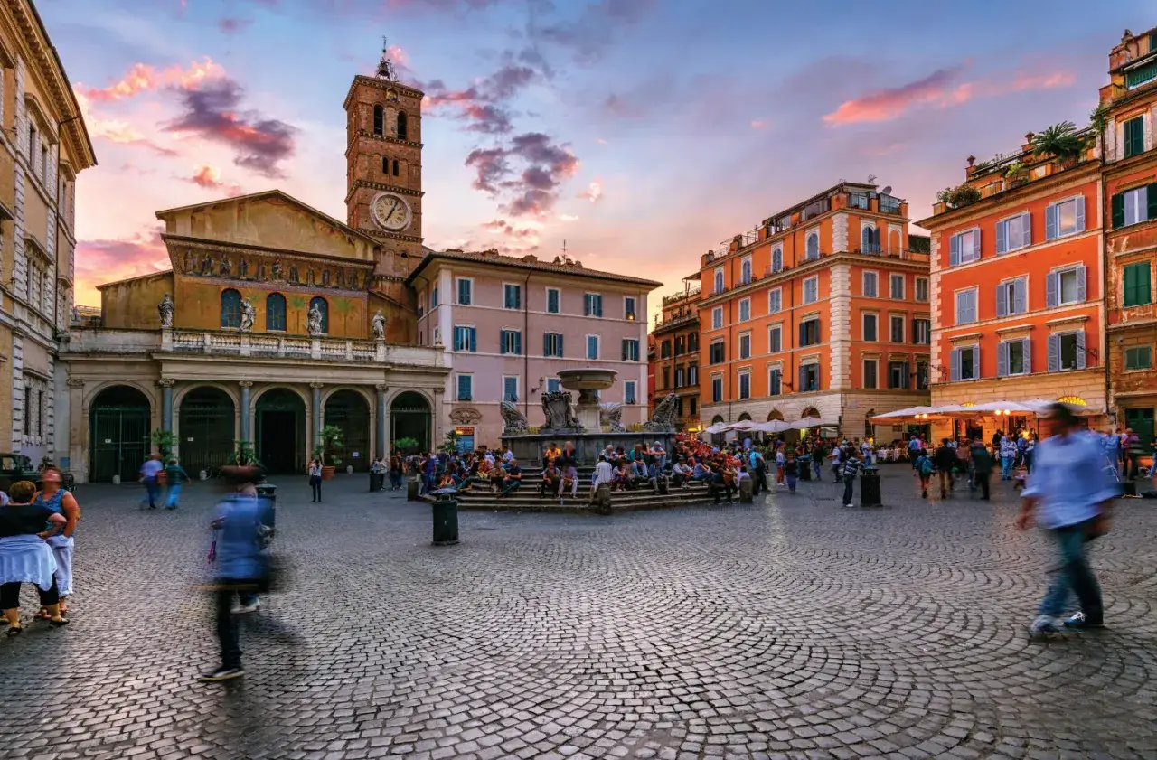 Wieczorny gwar na placu w Trastevere. Ludzie siedzą przy fontannie, a w tle widać kościół z wieżą i kolorowe kamienice.