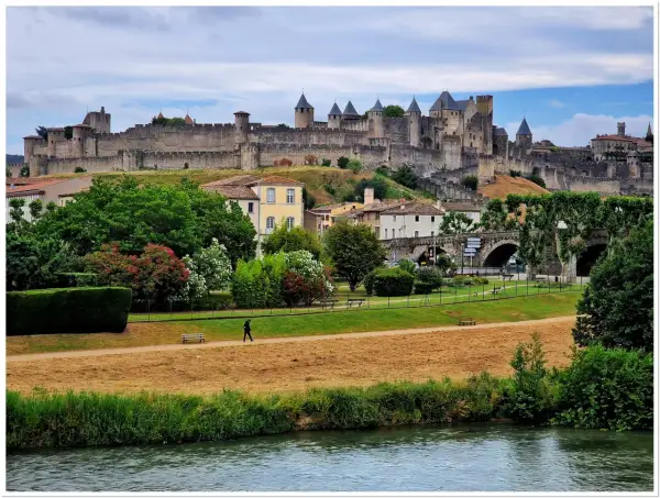 Majestatyczna twierdza Carcassonne góruje nad miastem. W dole zielony park, rzeka i spacerująca postać.
